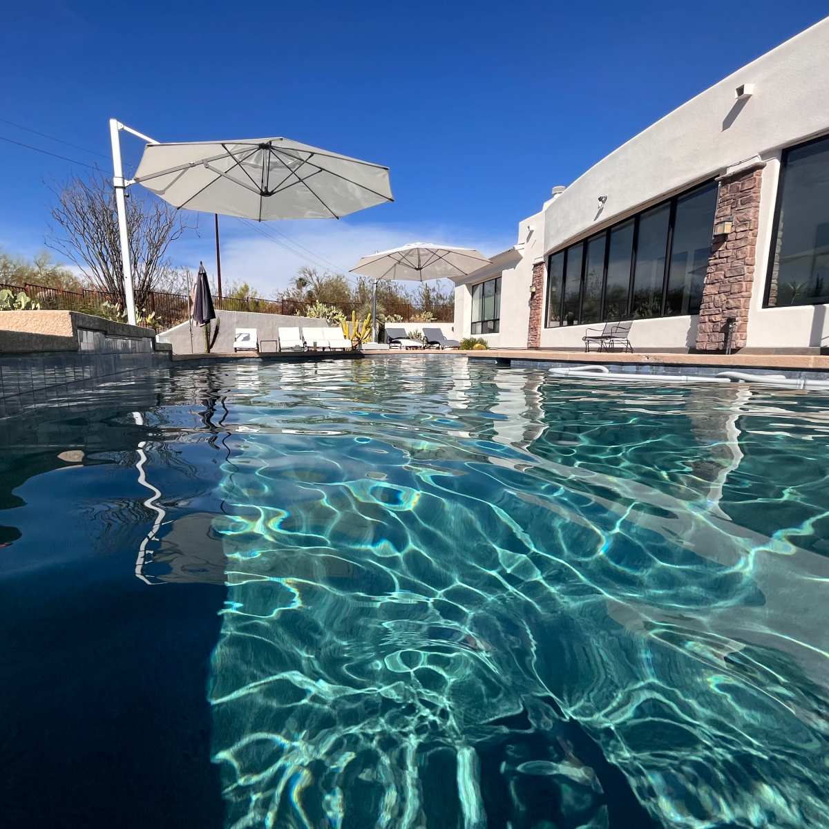A clear swimming pool with reflections of umbrellas and a house against a blue sky.