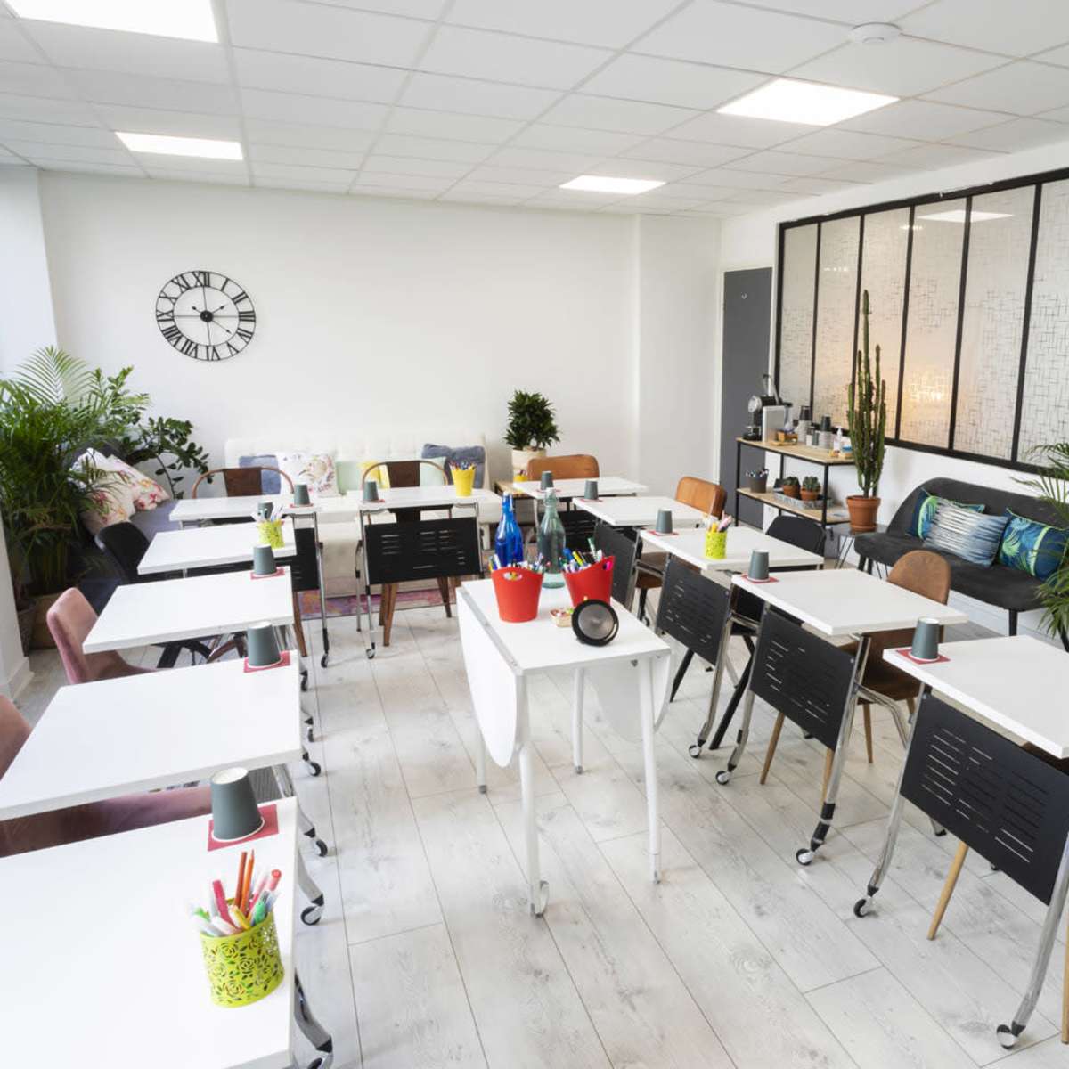 A modern classroom setup with multiple tables and chairs arranged neatly, surrounded by plants and featuring a wall with large windows.
