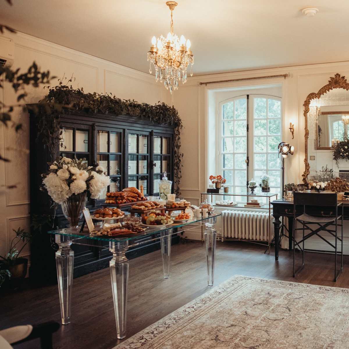 A elegantly decorated room featuring a table with assorted pastries, a mirror, and large windows allowing natural light, complemented by decorative chandeliers and greenery.