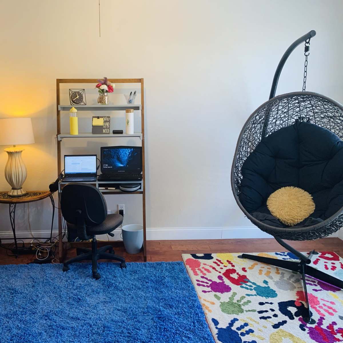 A living space featuring a black leather couch, a desk with a computer, and a hanging chair, all on a vibrant blue rug.