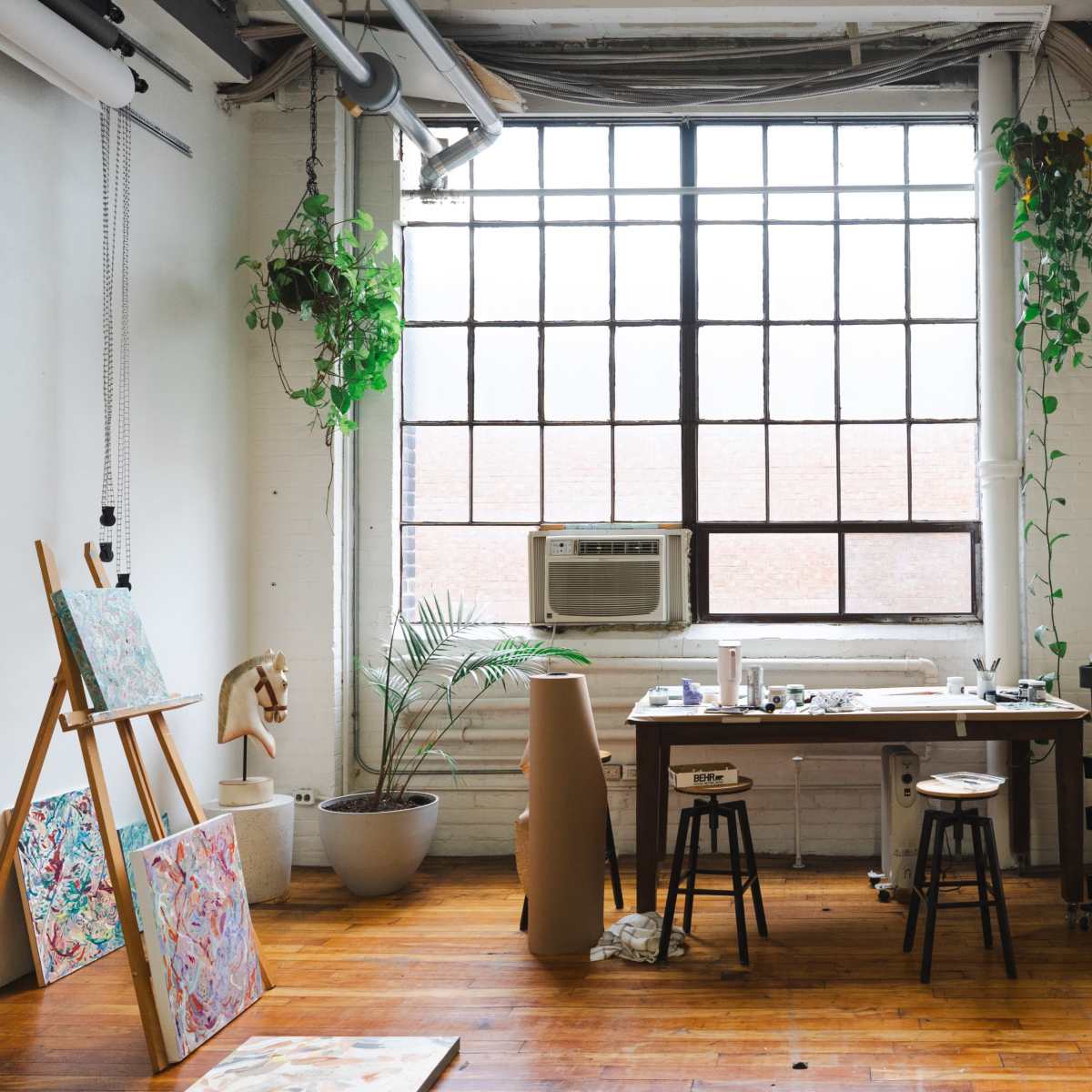 A bright art studio features large windows, various potted plants, a wooden table cluttered with art supplies, and easels displaying colorful artworks.