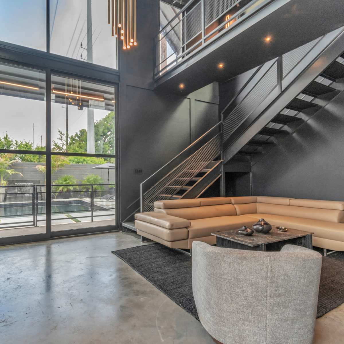 A modern living area featuring a light-colored sectional sofa, a glass wall providing outdoor views, and a staircase leading to an upper level.