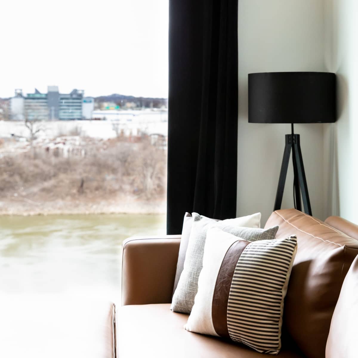 A brown couch with decorative pillows next to a floor lamp, overlooking a view through a large window.