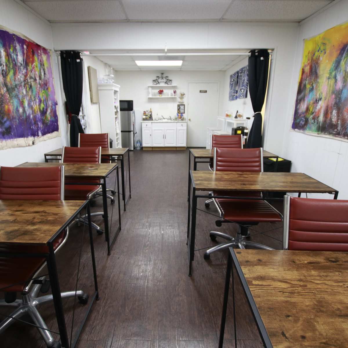 A spacious room with several wooden tables and chairs arranged in two rows, leading to a kitchenette area at the back, adorned with colorful artwork on the walls.