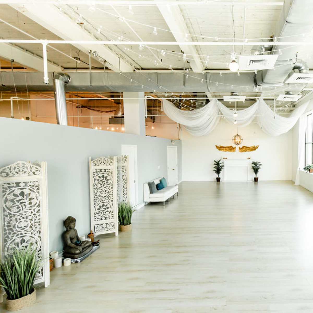 The spacious interior features light-colored wooden flooring, large windows with sheer curtains, and minimal decor including a Buddha statue and ornamental screens.