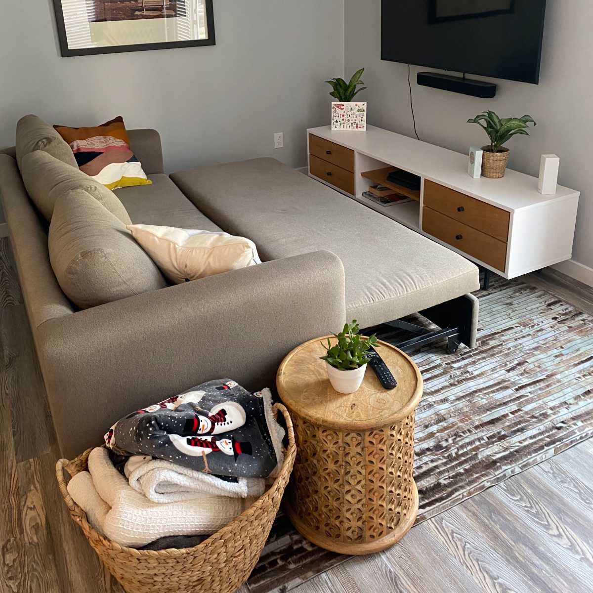 A cozy living room features a sectional sofa, a mounted TV, and a coffee table with decorative plants and storage baskets.