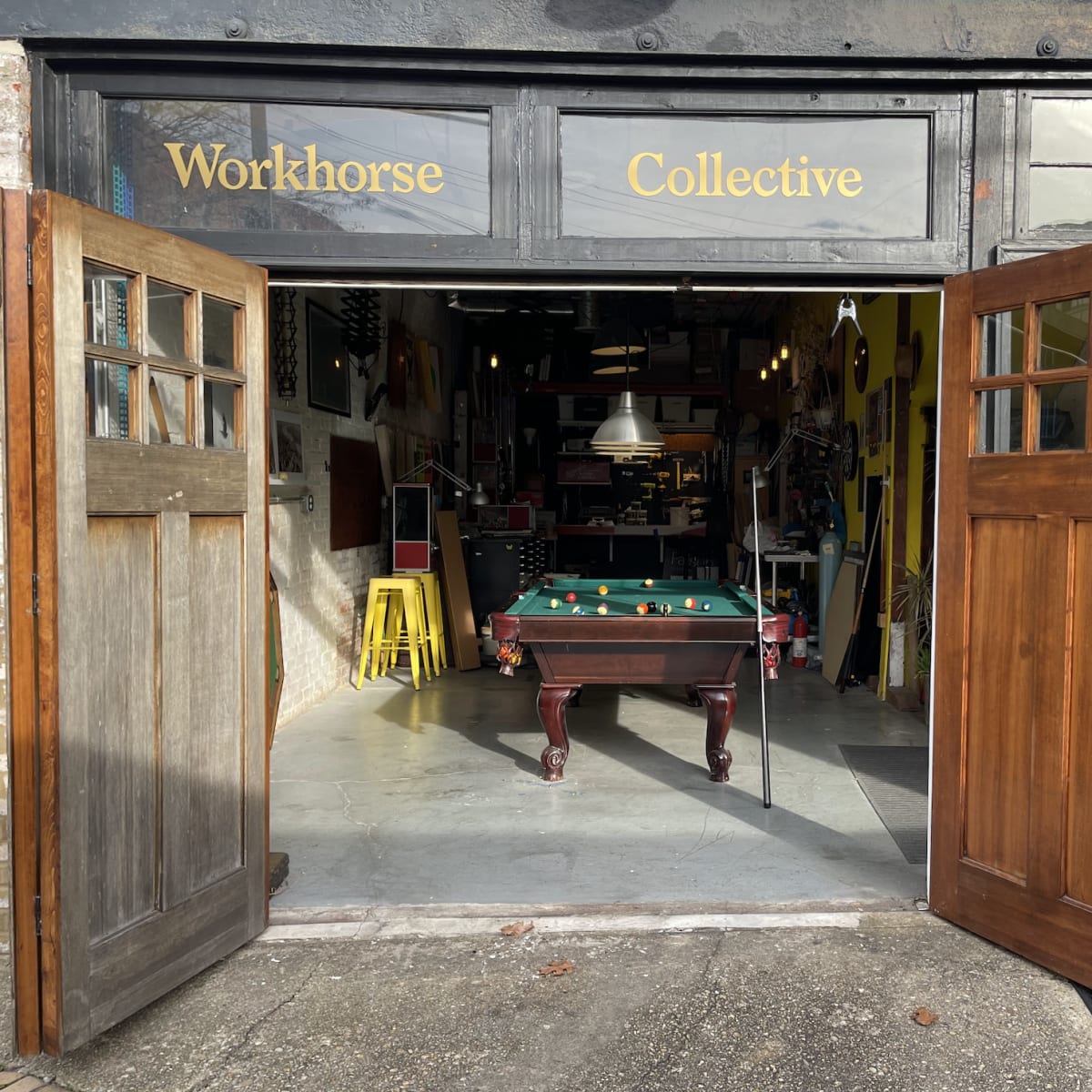 The entrance of a workshop named "Workhorse Collective" features an open double door leading to an interior with a green pool table and various tools and equipment visible inside.