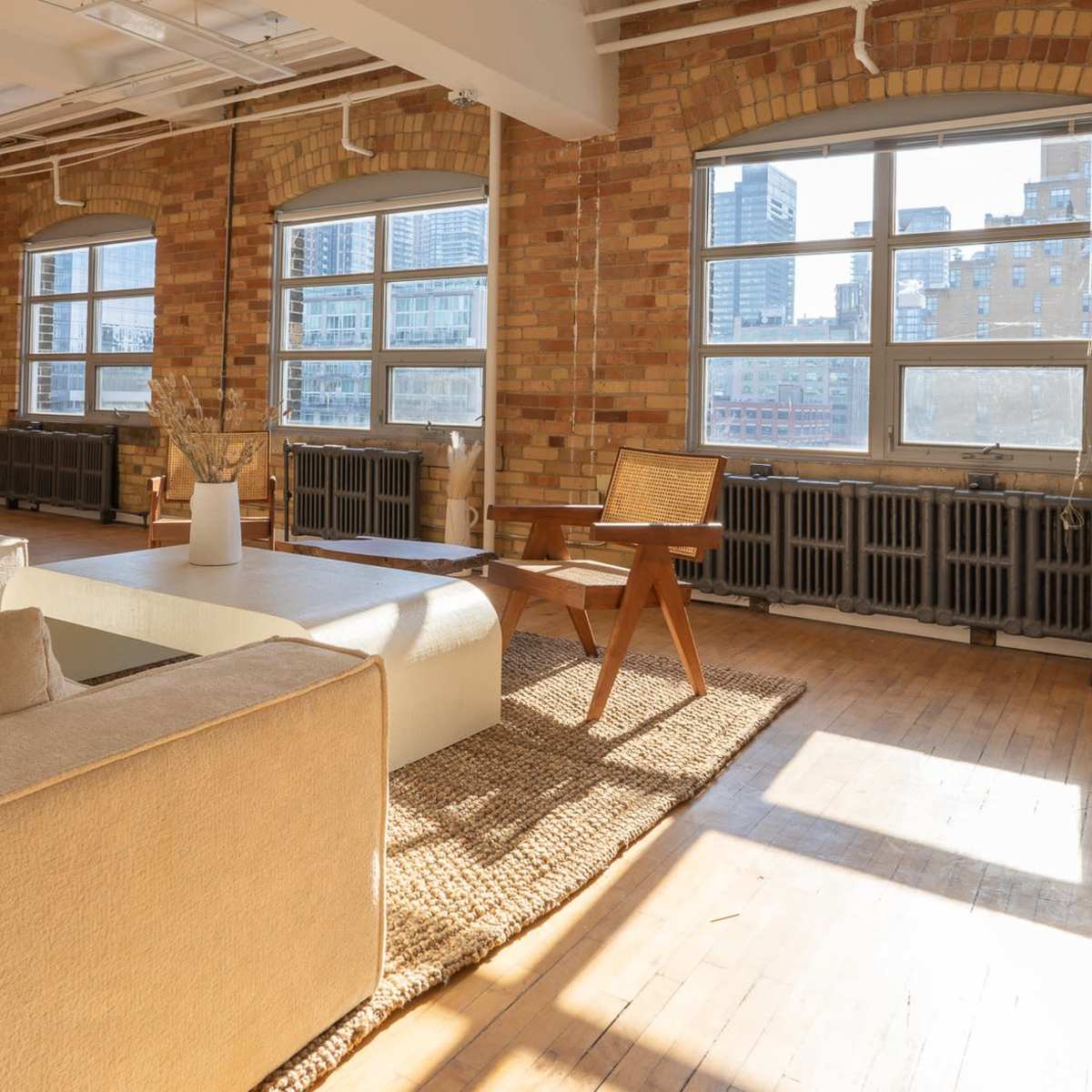 A spacious, sunlit living room features large windows, a beige sectional sofa, a wooden chair, and a large mirror against a brick wall.