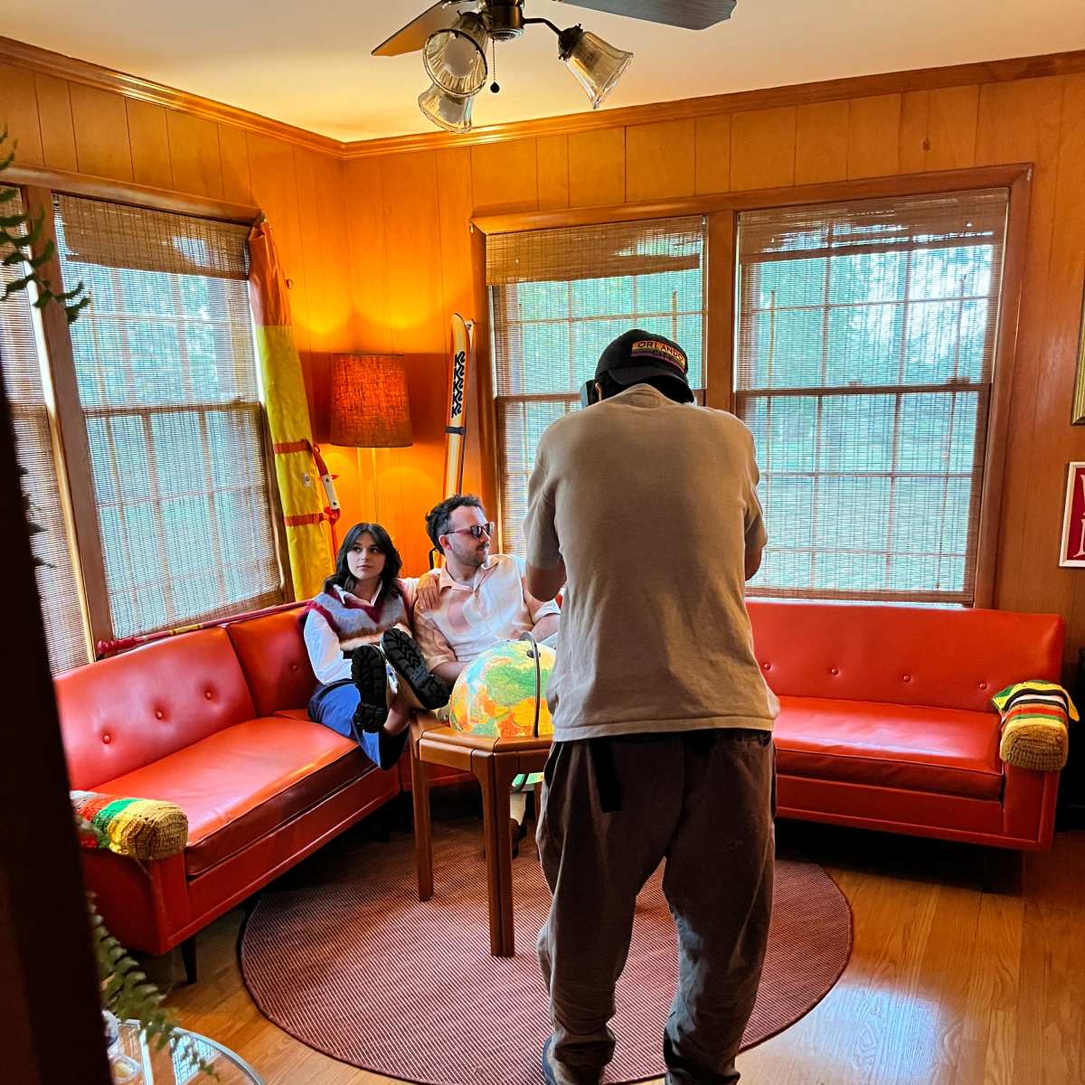 A person takes a photo of a couple sitting on a round table in a living room with large windows and red sofas.