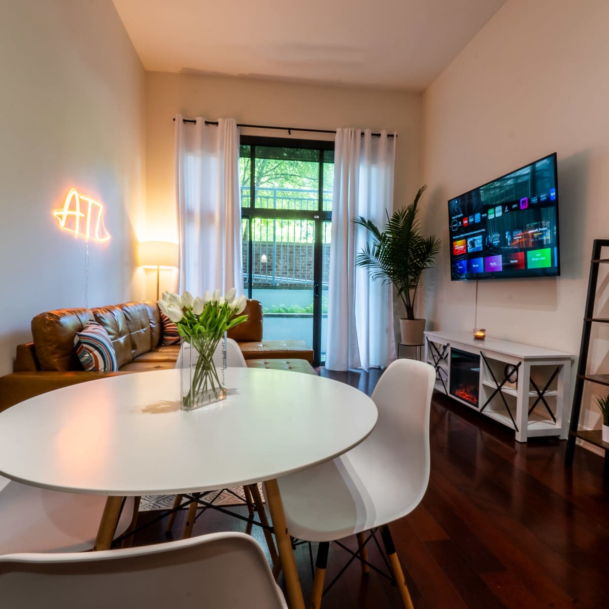 A cozy living area features a sofa with decorative pillows, a round dining table with white chairs, and a television on a stand, complemented by a window with sheer curtains and a view of an outdoor space.