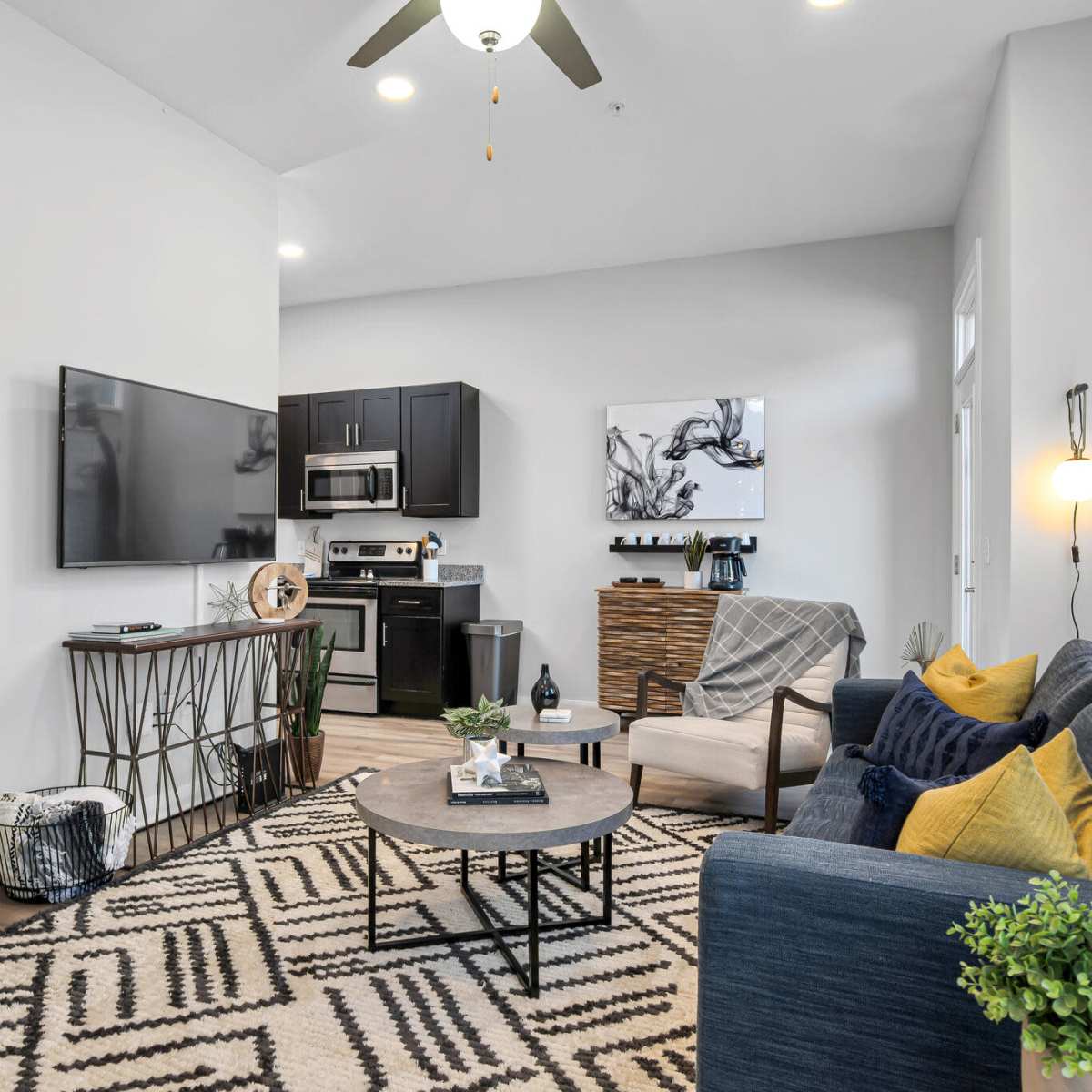 A modern living space featuring a kitchen area with dark cabinetry, a television mounted on the wall, a round coffee table, and seating arrangements that include a blue sofa and a patterned rug.