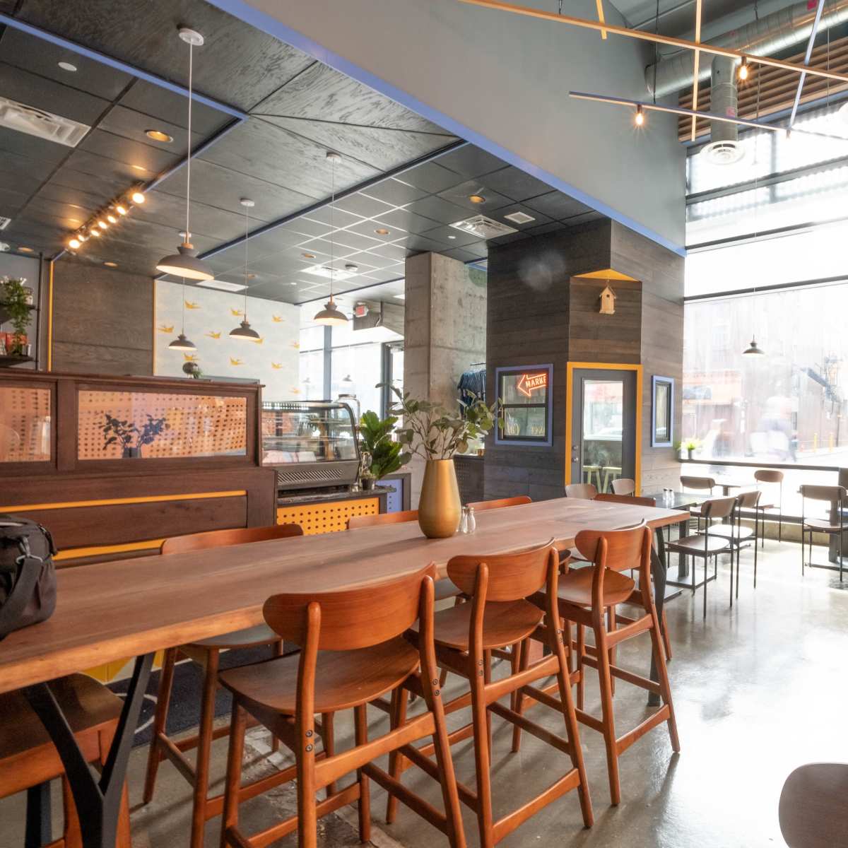 A modern restaurant interior with wooden tables, high-backed chairs, and large windows allowing natural light to enter.