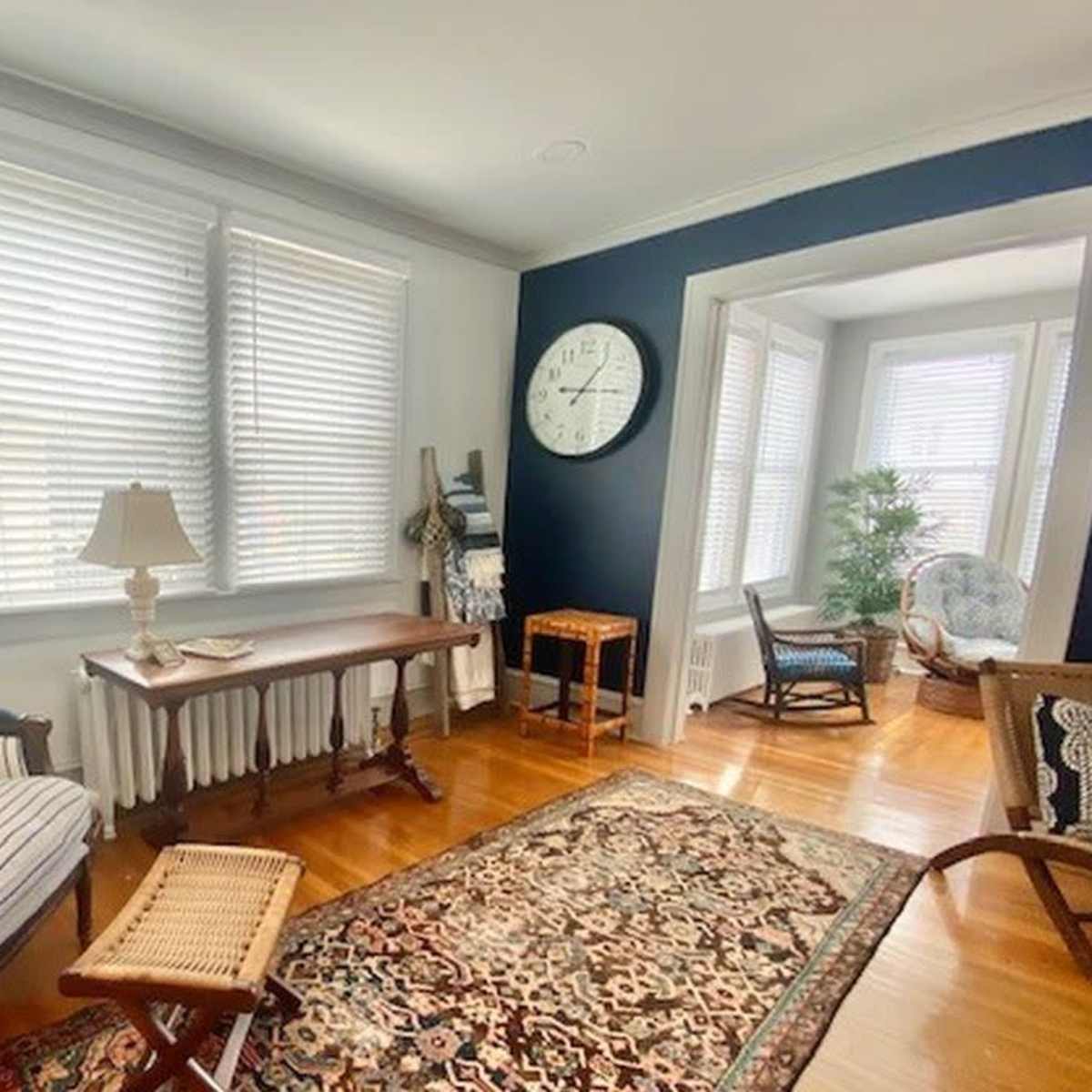 The room features a striped chair with a lamp on a wooden table, a large wall clock, and a decorative area rug on hardwood floors.