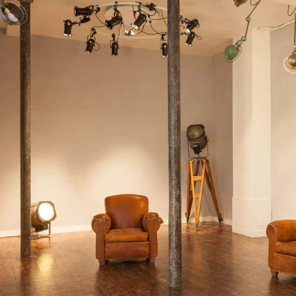 A minimalist interior space featuring two brown leather armchairs, a tall wooden tripod light stand, and various mounted lights on the ceiling.