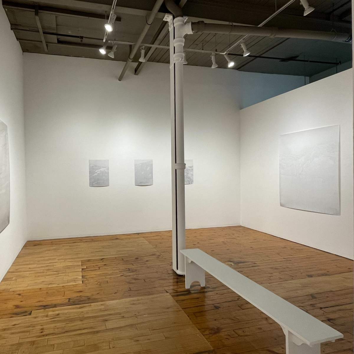 An art gallery with white walls, wooden flooring, and a white bench in the center, featuring several minimalist artworks displayed on the walls.
