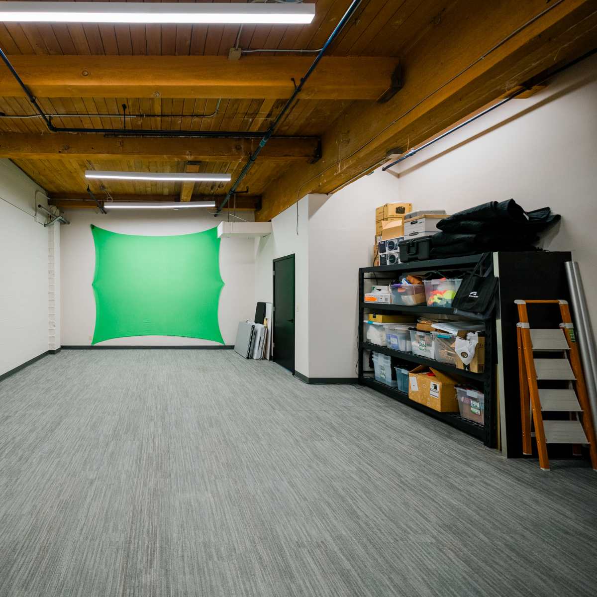 A large, empty room with wooden beams, a green backdrop on the wall, and storage shelves filled with various materials along one side.