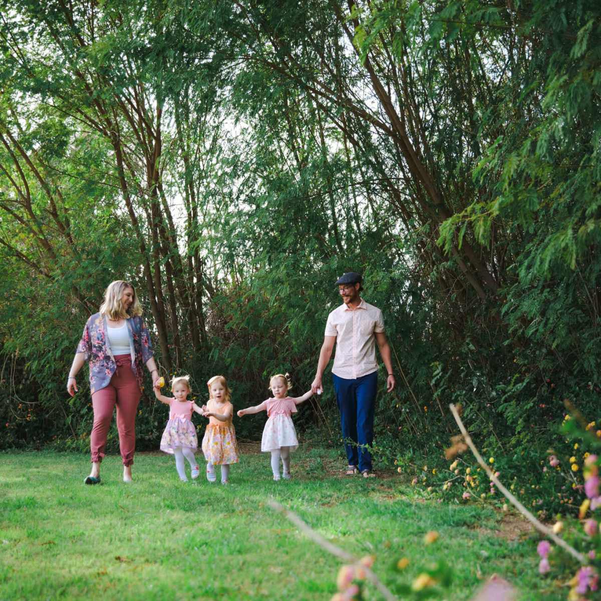 A woman and a man walk hand-in-hand with three young girls through a grassy area surrounded by trees.
