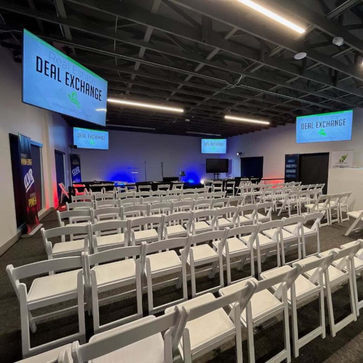 A conference room setup with multiple rows of white chairs facing a stage area lit with blue and red lighting, and two large screens displaying "DEAL EXCHANGE."