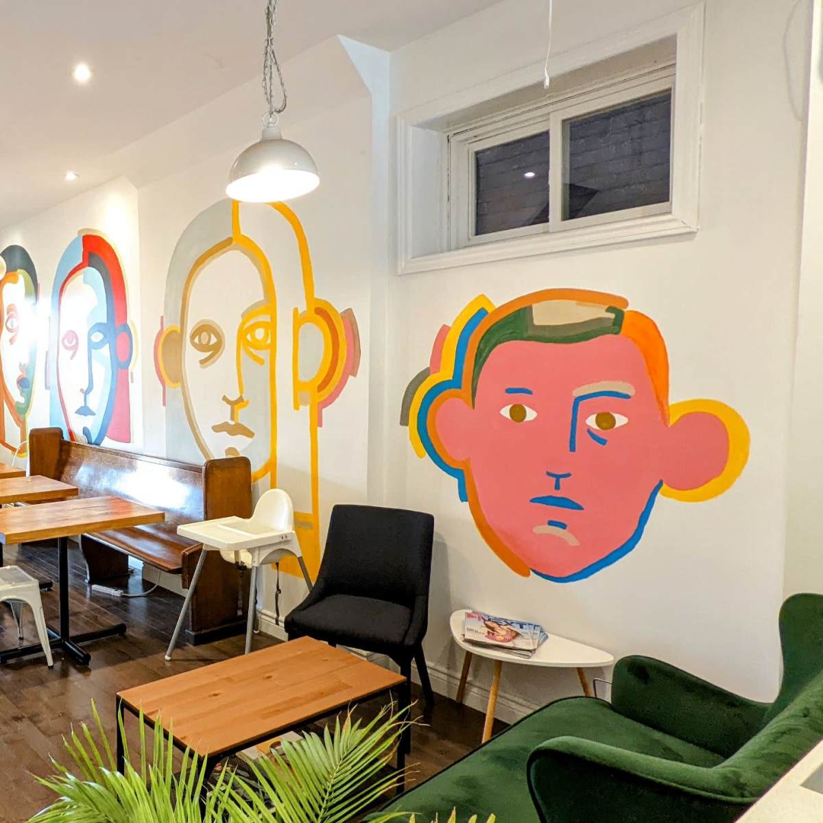 A modern café interior featuring wooden tables, a staircase, and colorful mural artwork of stylized faces on the walls.