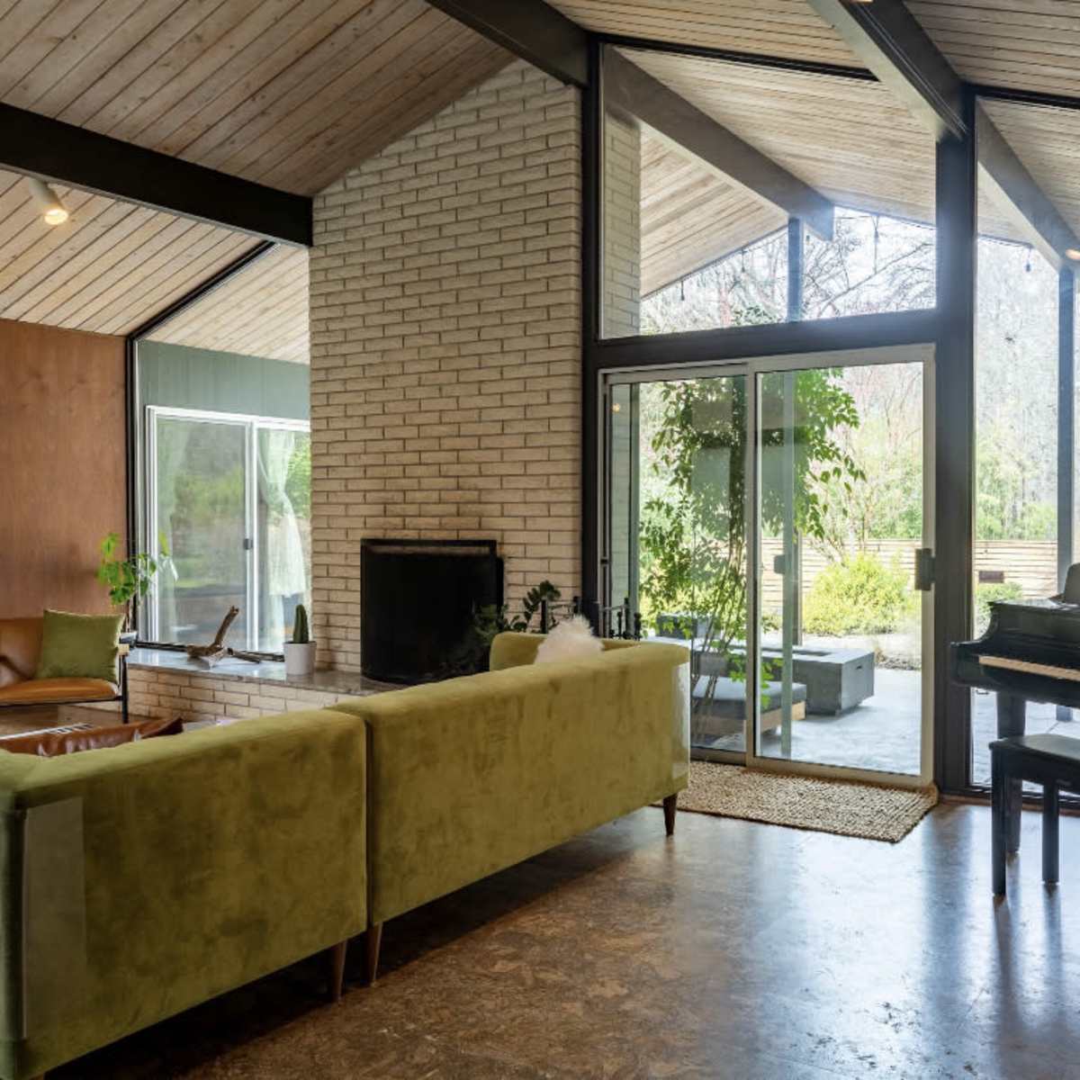 A spacious living room features a green sofa, a black piano, and large glass doors that open to a landscaped outdoor area.