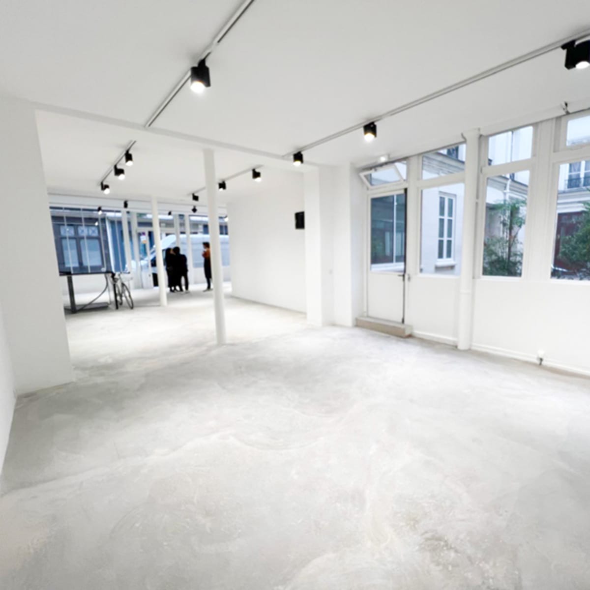 A spacious, empty room with concrete flooring and large windows, illuminated by overhead lighting.