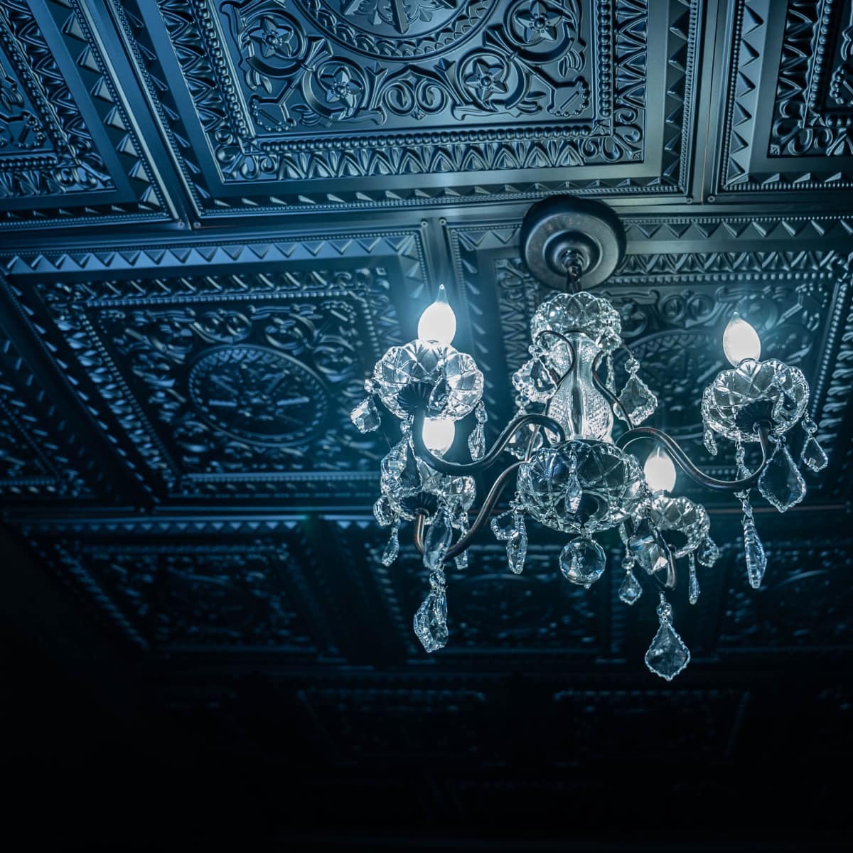 A crystal chandelier with multiple light bulbs hangs from a decorative, intricately patterned ceiling.