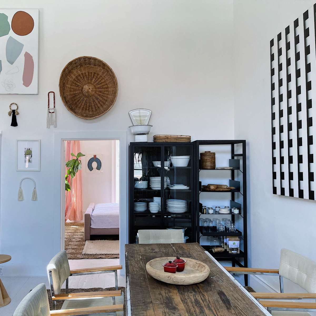 A bright dining area features a wooden table, chairs, and various storage solutions, with art on the walls and an open space leading to a bedroom.