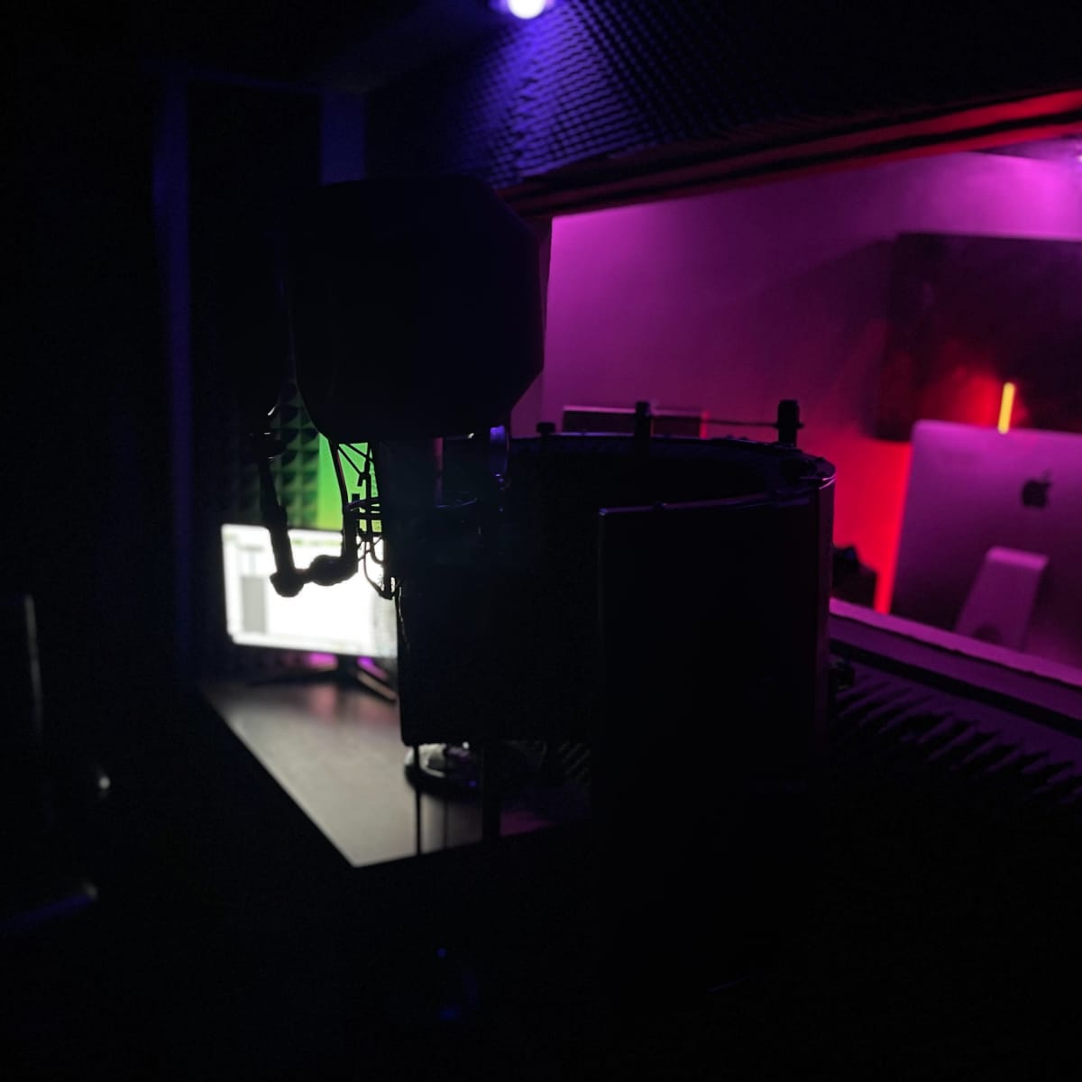 A microphone stands in silhouette against a dimly lit recording studio featuring colorful LED lights and computer screens.