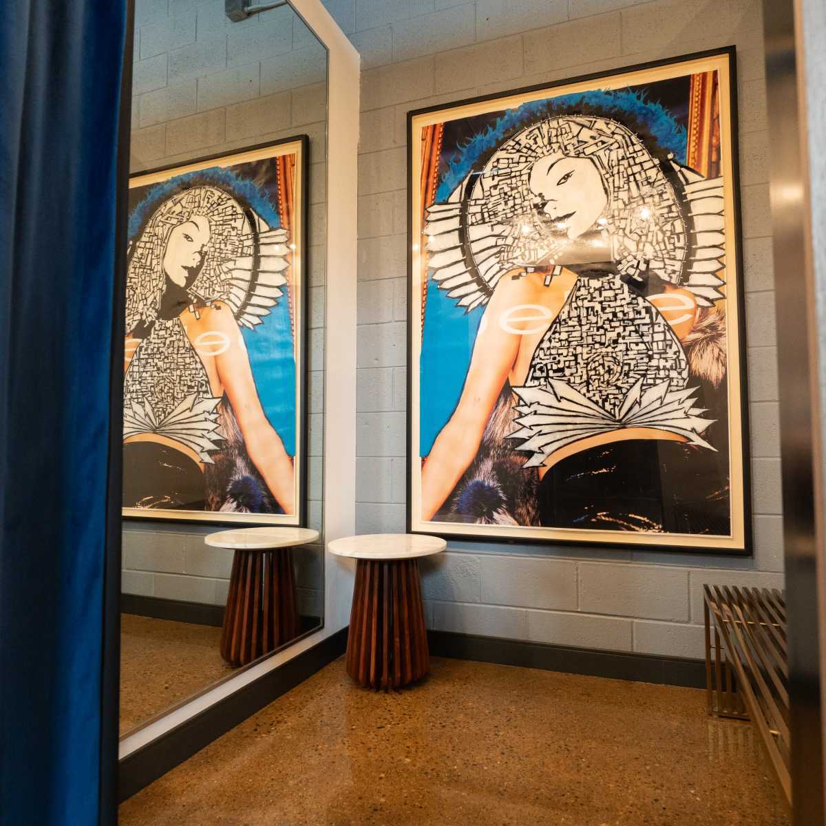 A room with two large, framed artwork pieces of a woman, flanked by a mirror and two small side tables, with a blue curtain hanging to one side.