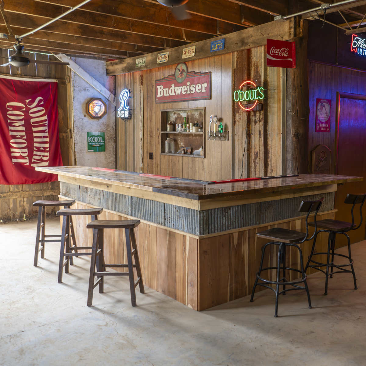 A rustic bar area with a wooden counter, several bar stools, and neon signs, including Budweiser and Southern Comfort, in a dimly lit room featuring wooden walls.