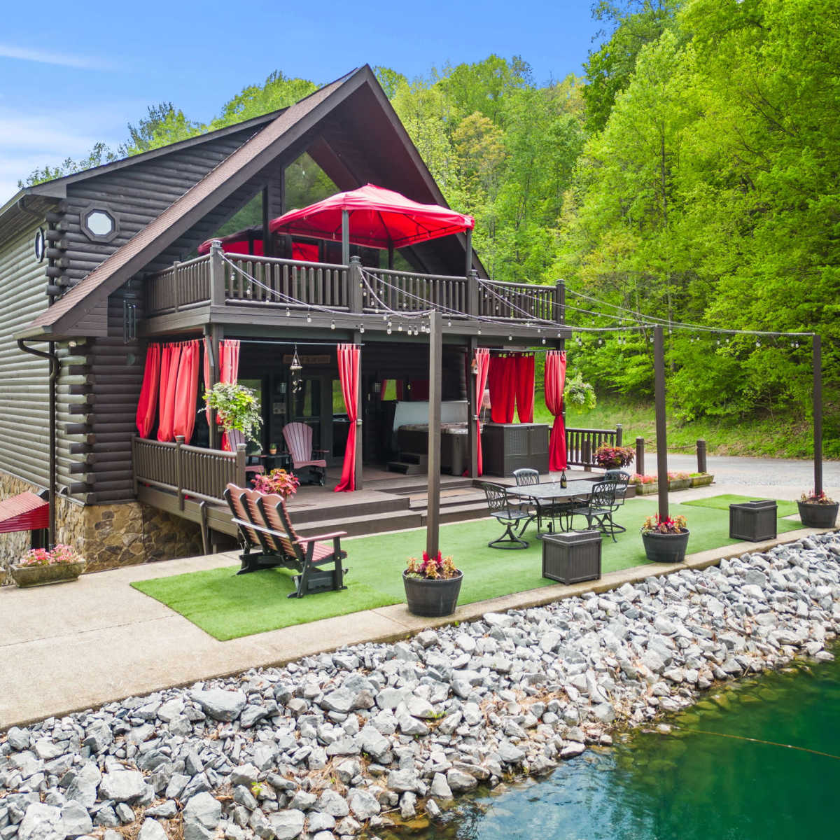 A two-story cabin with a large deck is situated beside a rocky shore and a calm body of water, surrounded by lush green trees.