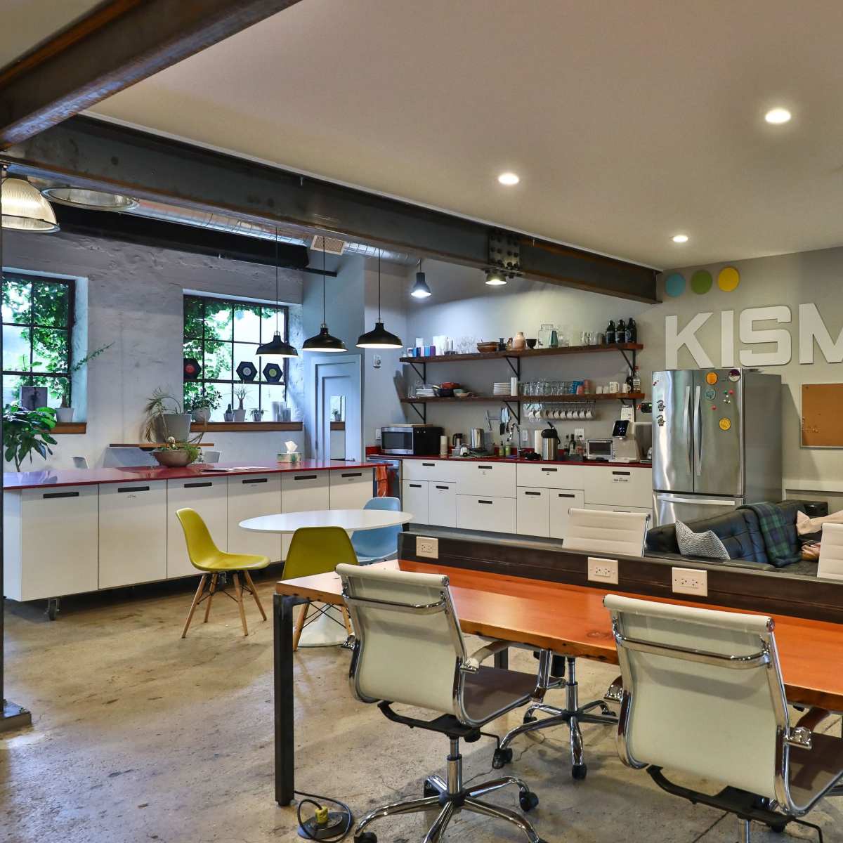 A modern office kitchen area featuring a communal table, chairs, a refrigerator, and shelves with various kitchen supplies, all adorned with the word "KISMET" on the wall.