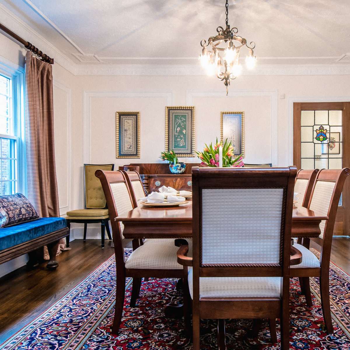 A dining room features a wooden table surrounded by eight chairs, a floral centerpiece, and vintage decor on the walls.