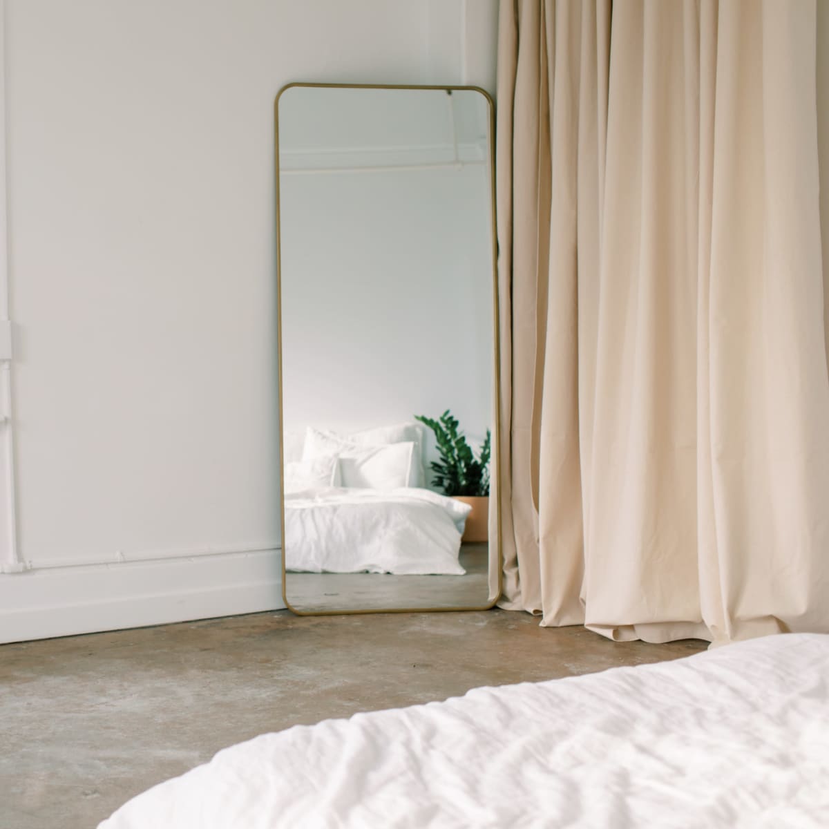 A minimalist bedroom with a large mirror next to a window, and a bed with white linens in the foreground.