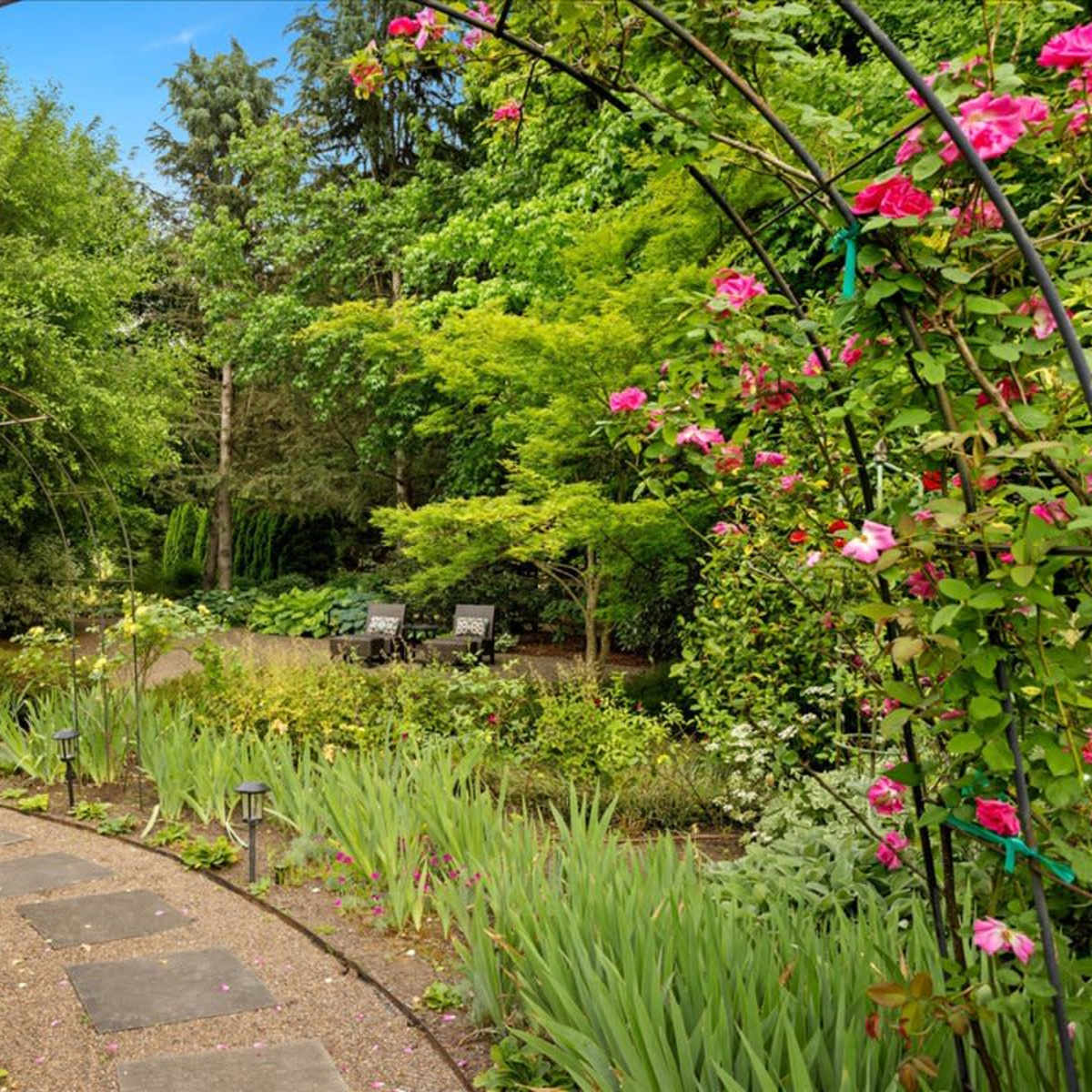 A stone pathway winds through a lush garden archway adorned with colorful flowers, leading to a secluded seating area.