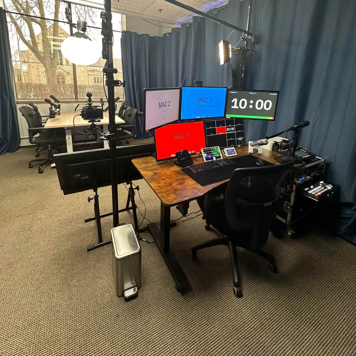 A multimedia production studio equipped with multiple screens, a control panel, and camera setups, surrounded by dark curtains in a room with large windows.