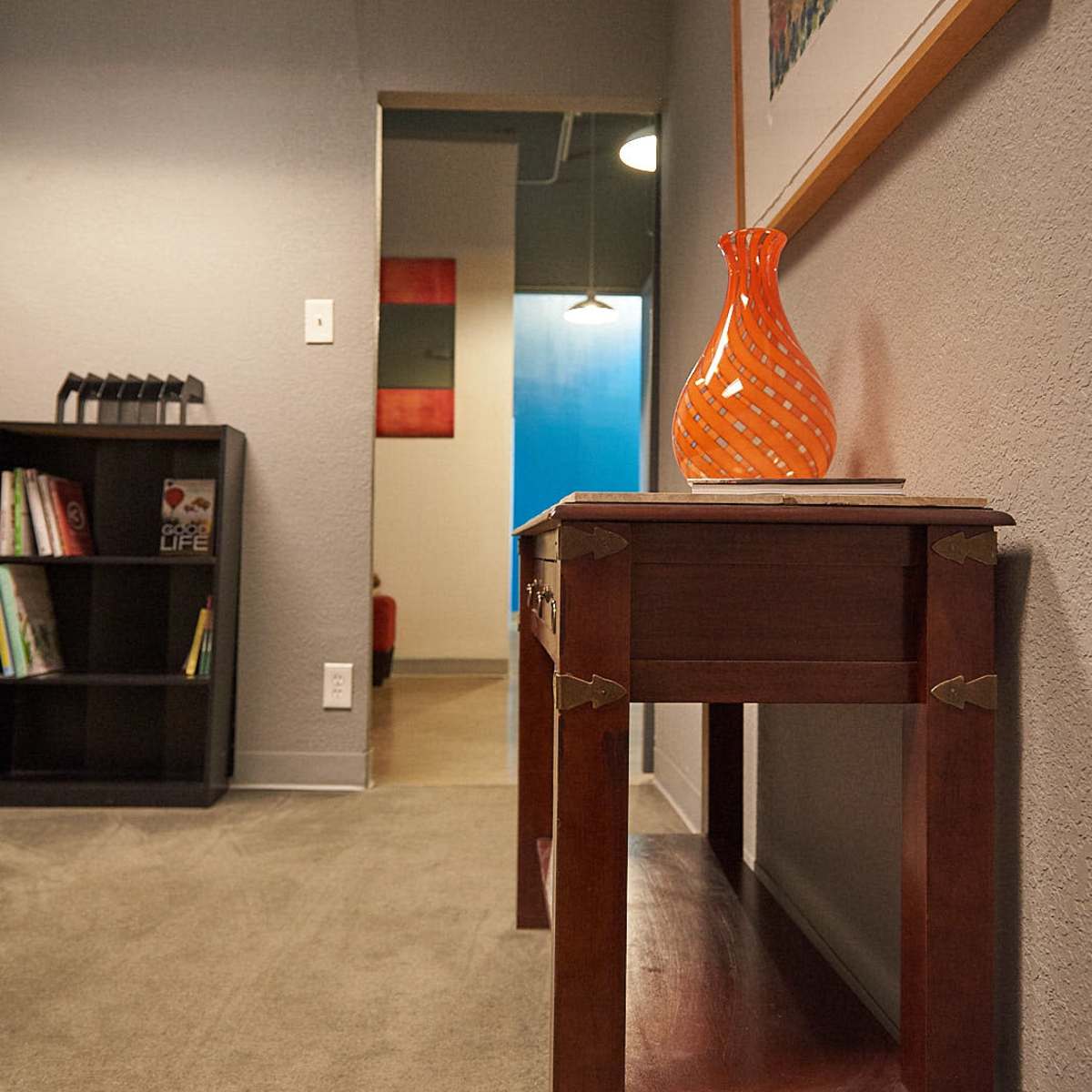 A wooden table with an orange vase on top is positioned in a hallway leading to rooms with colorful artwork.