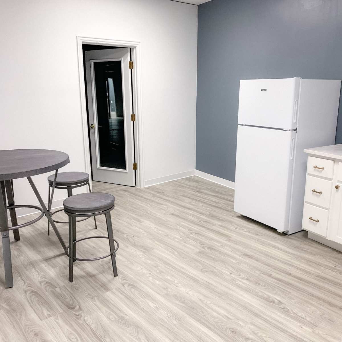 A simple kitchen area featuring a white refrigerator, a round dining table with stools, and a gray wall.