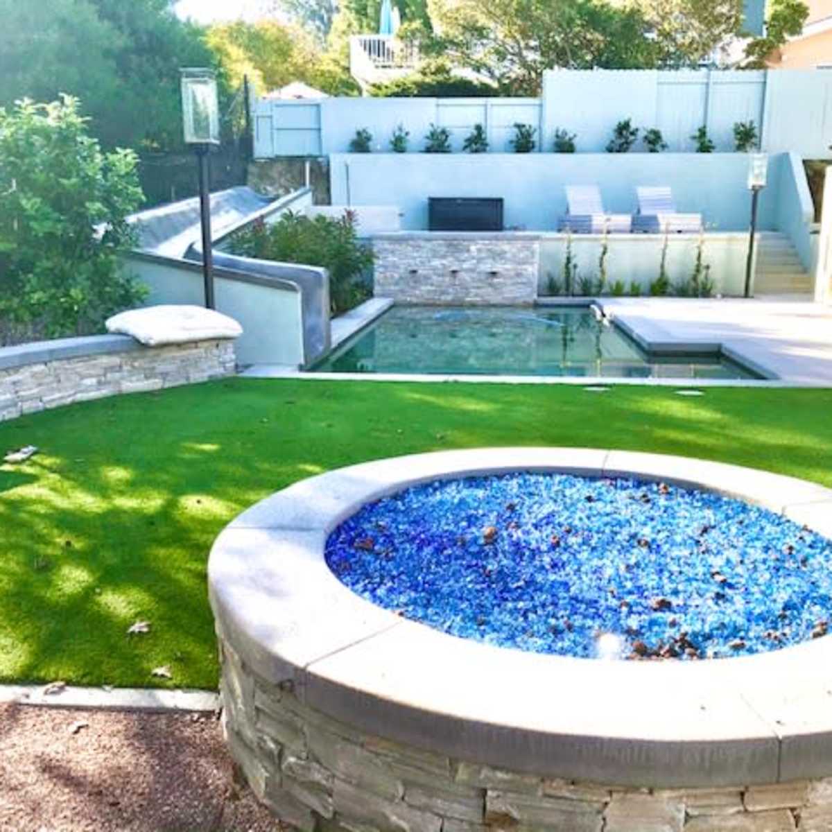 A landscaped backyard featuring a round fire pit surrounded by blue glass, a green lawn, and a swimming pool with a stone backdrop and lounge chairs.