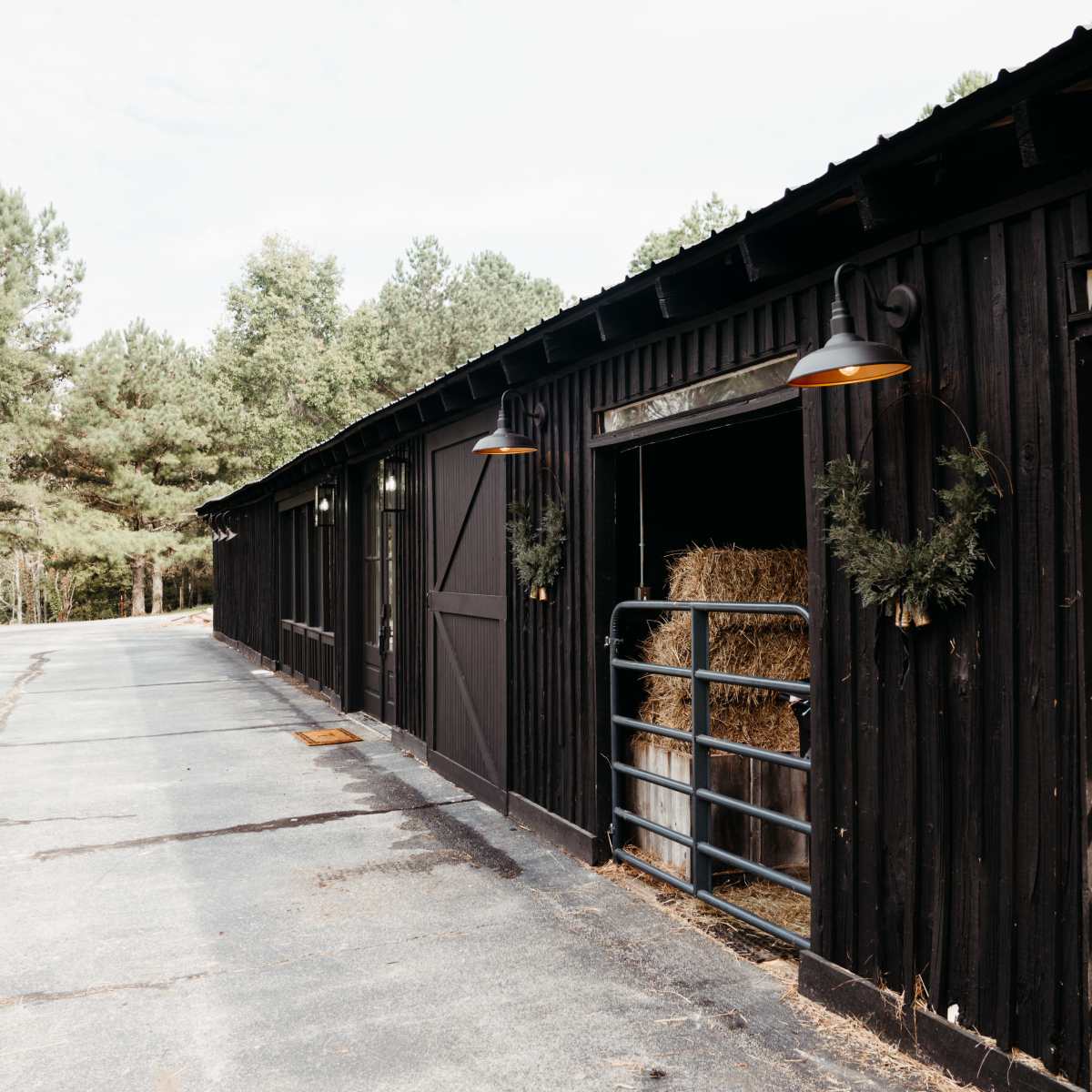 A black barn with open stalls displays bales of hay, lined along a paved path surrounded by trees.