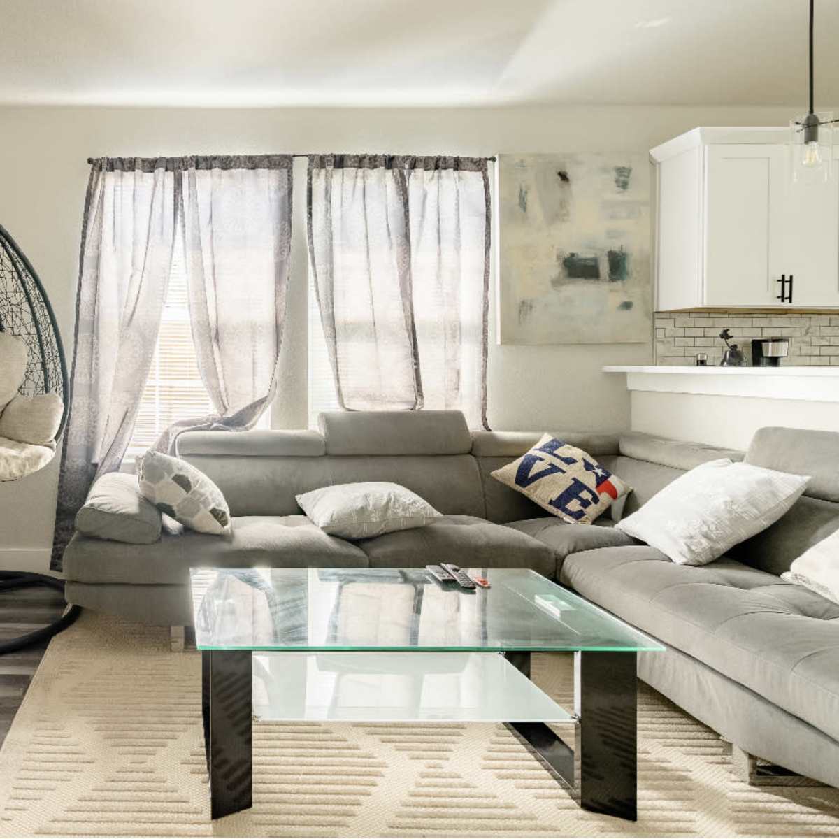 A modern living room with a gray sectional sofa, a glass coffee table, and a hanging chair beside large windows with light curtains.