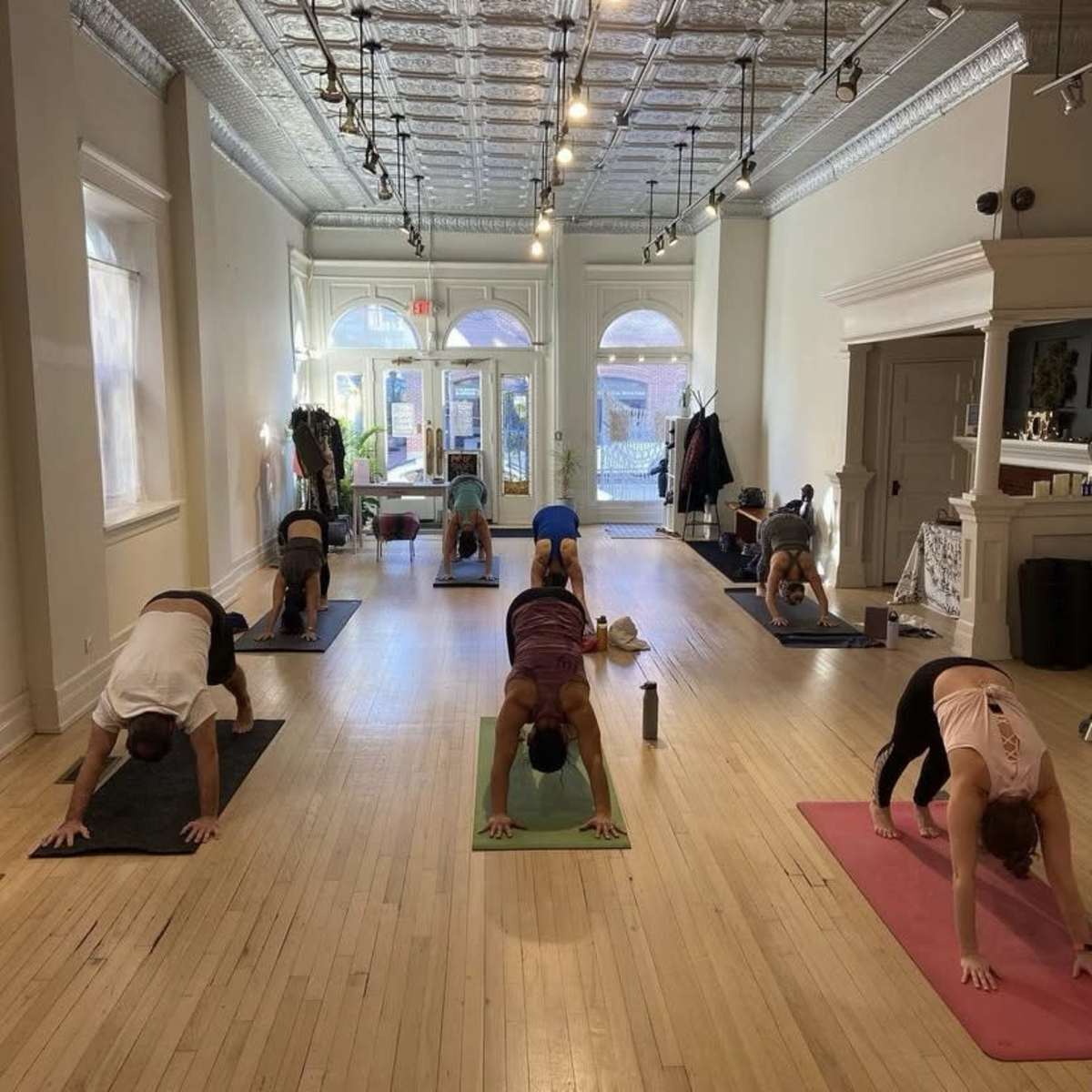 Several people participate in a yoga class, practicing downward dog poses on mats in a well-lit studio with wooden floors.