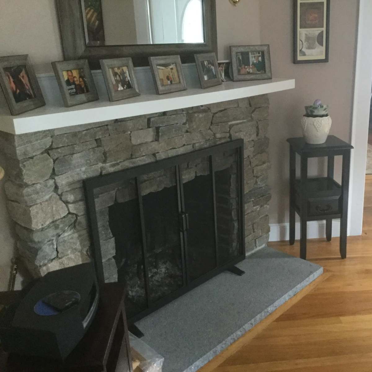 A living room with a stone fireplace, a wooden floor, framed pictures on the mantel, and a small table beside the fireplace.