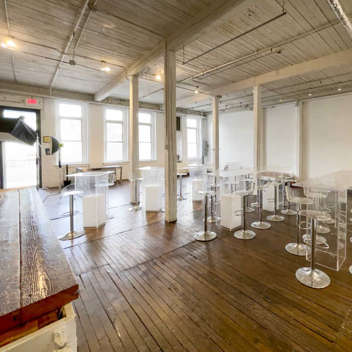 A spacious room with wooden floors, featuring transparent acrylic tables and stools arranged in a modern layout, alongside a few white seating areas.