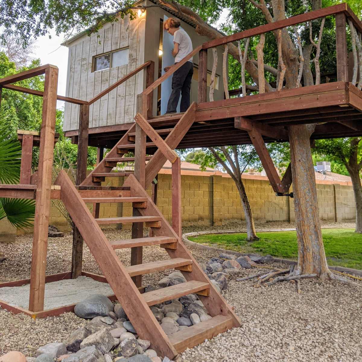 A person is using a light fixture on the exterior of a wooden treehouse elevated above a set of stairs in a landscaped yard.