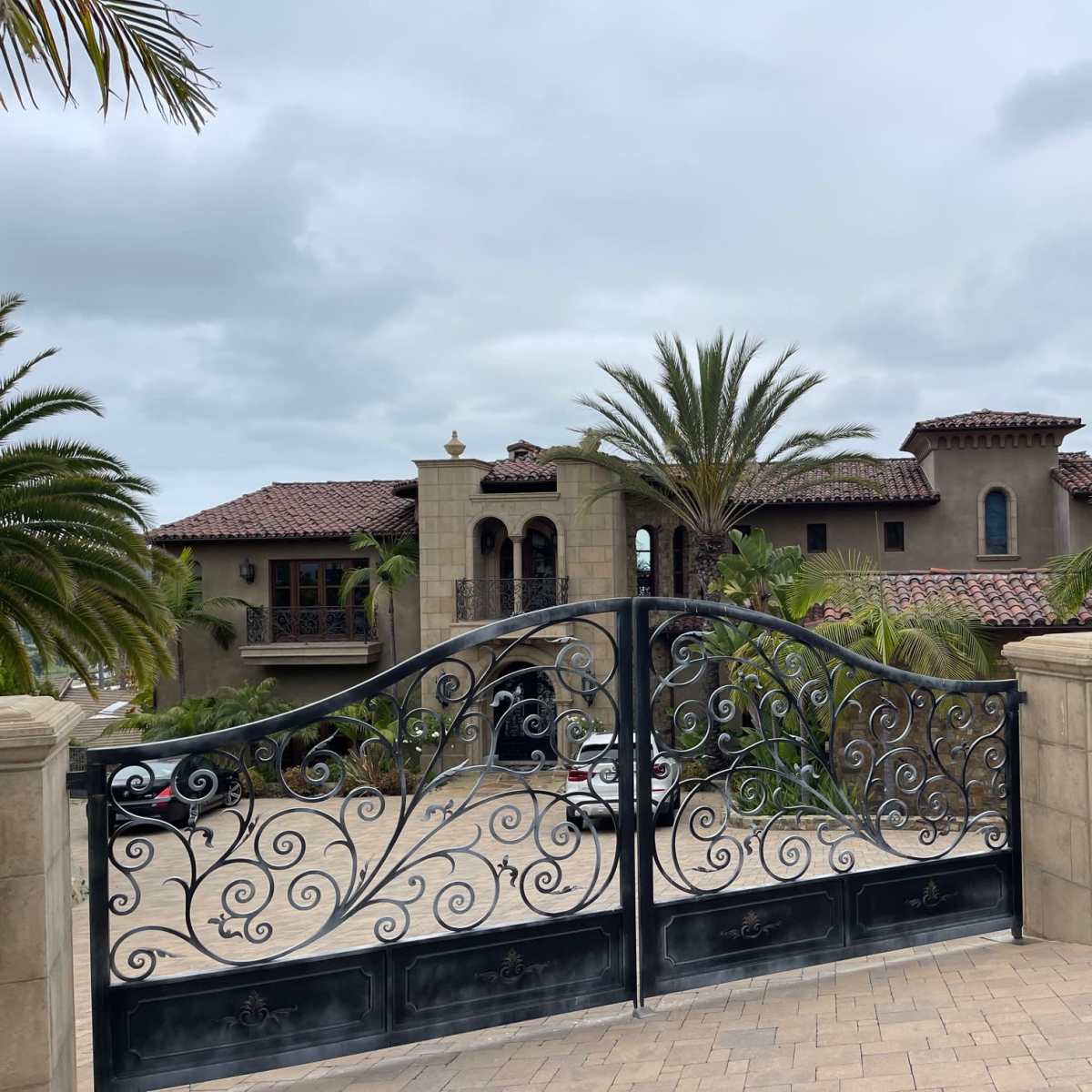 A lavish stone mansion behind ornate black gates, surrounded by palm trees and lush greenery under a cloudy sky.
