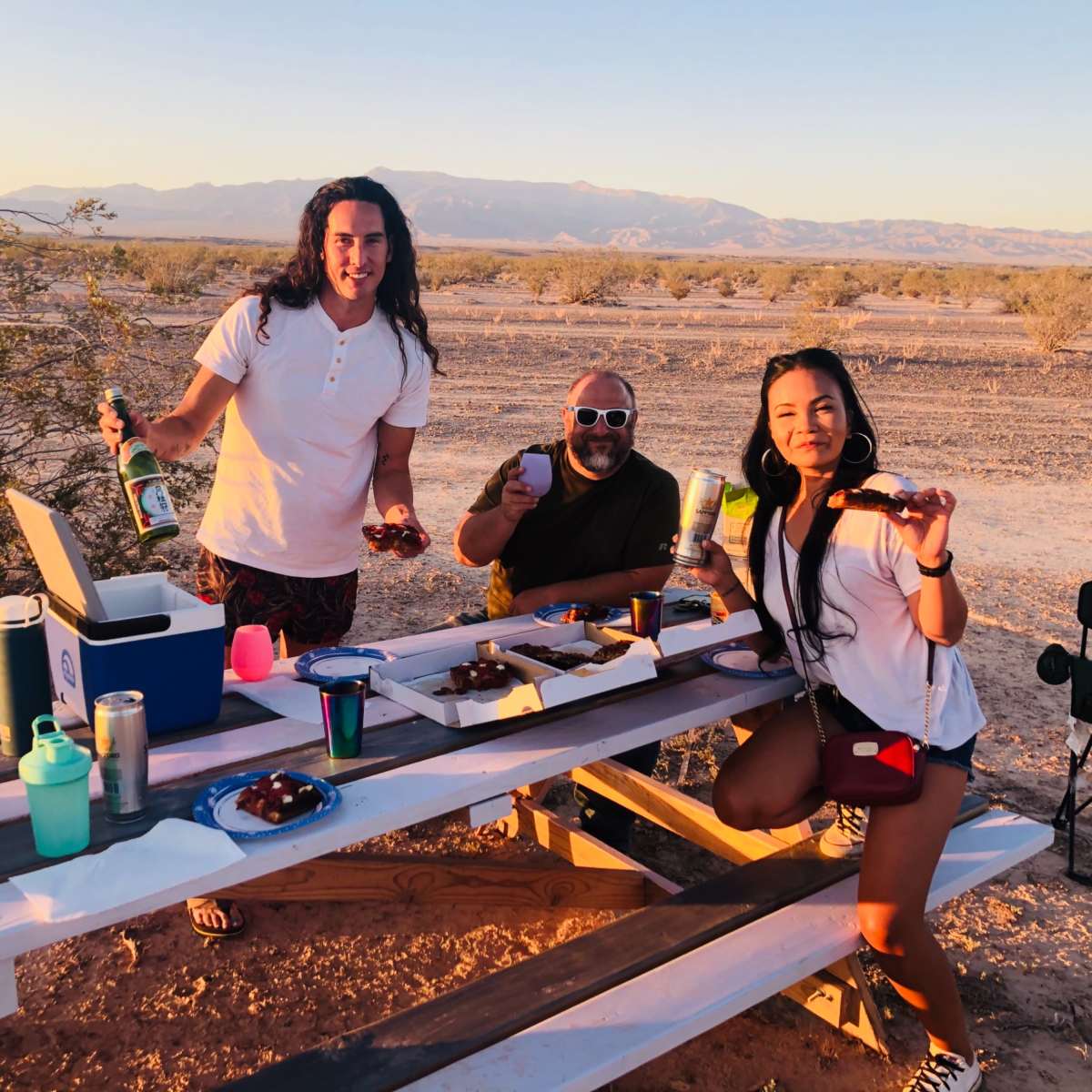 Three people hold drinks and snacks while posing at a picnic table in a desert landscape during sunset.