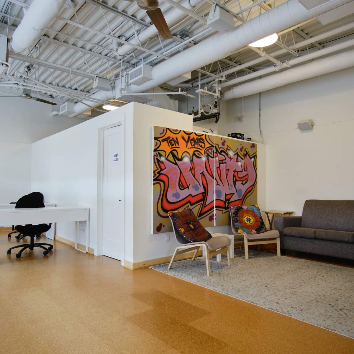 A modern office space features a gray sofa, two chairs, a decorative rug, and a wall with colorful graffiti art.