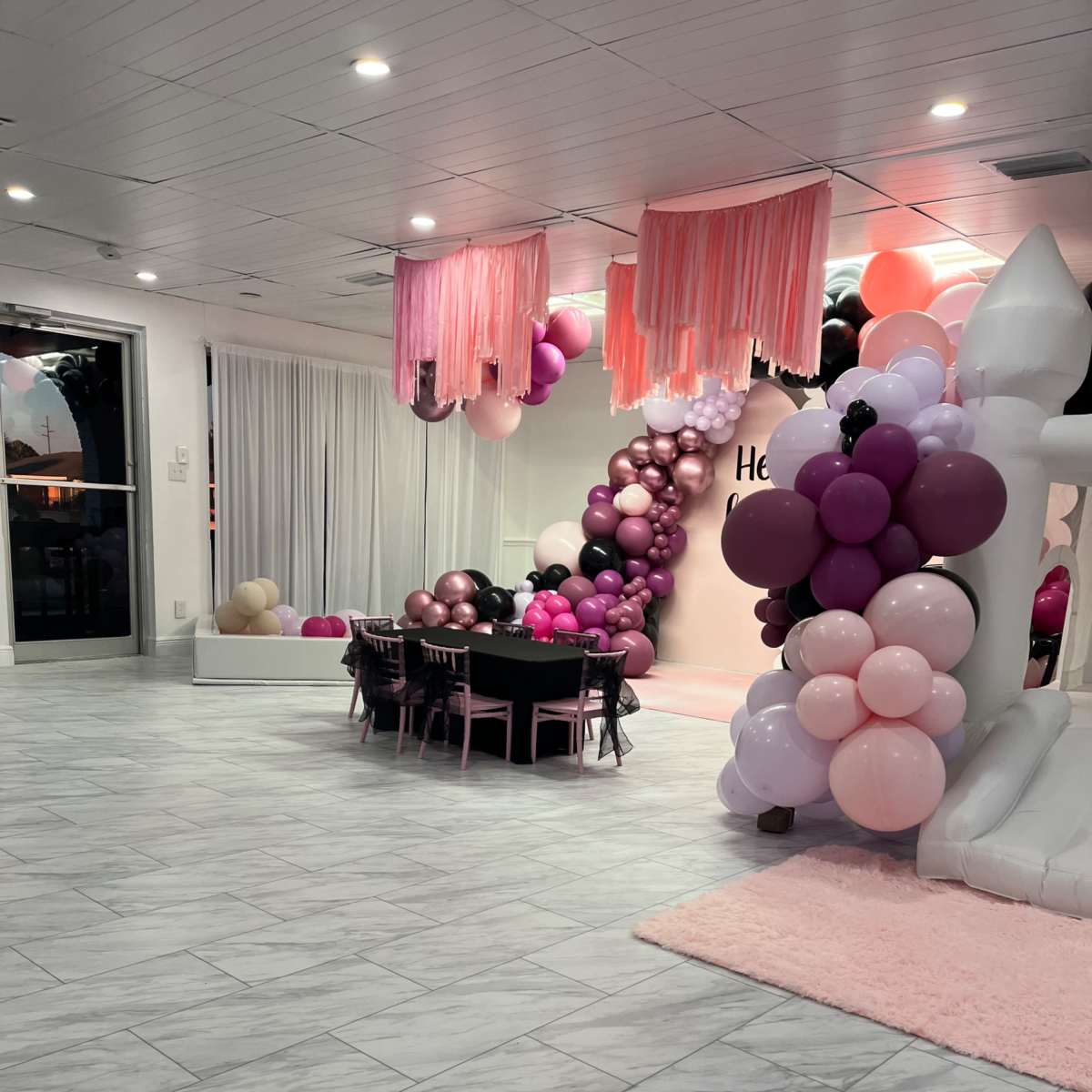 The scene features a spacious event room decorated with pink, black, and purple balloons, a table set with chairs, and a white bounce house in one corner.