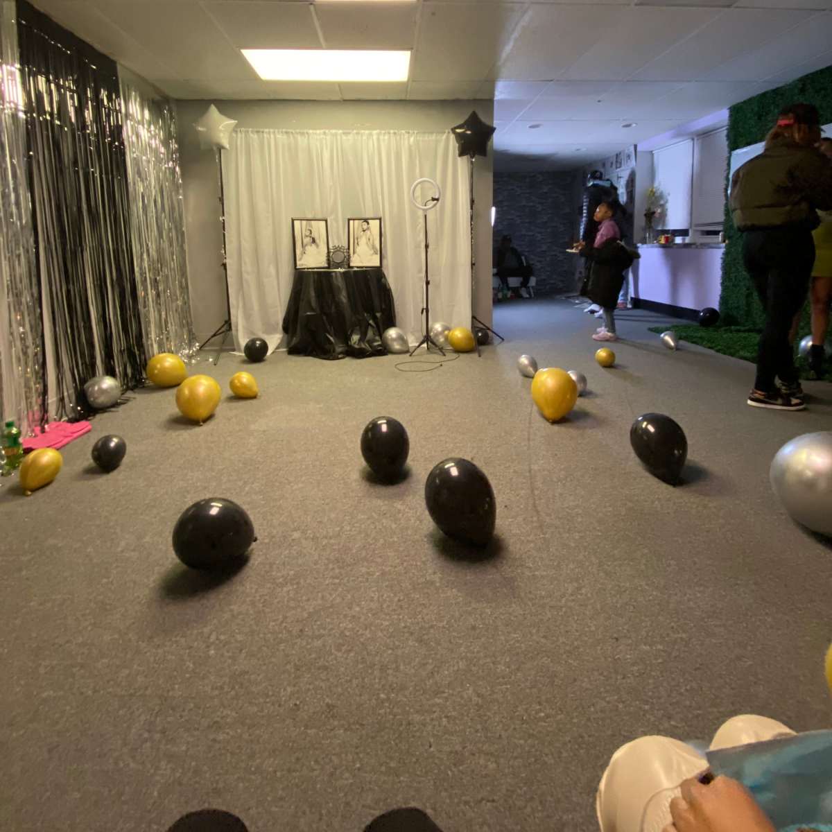 A room decorated with black and gold balloons scattered across the floor, featuring a backdrop with a small setup for a photo display at the far end.